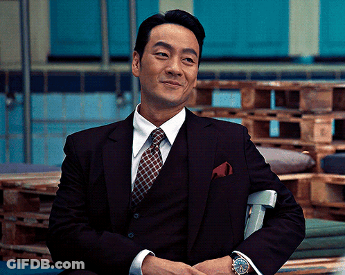 A Man In A Suit And Tie Sits In A Chair With His Hands Folded GIF