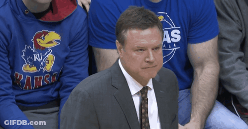 A Man In A Suit And Tie Sits In Front Of A Kansas Sweatshirt GIF