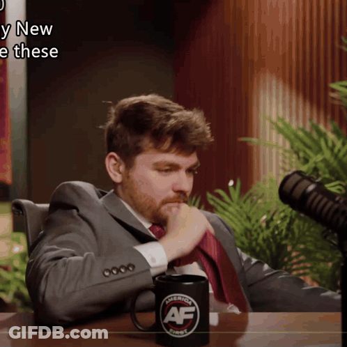 A Man In A Suit And Tie Sits In Front Of A Microphone With A Mug That Says " America First " On It GIF