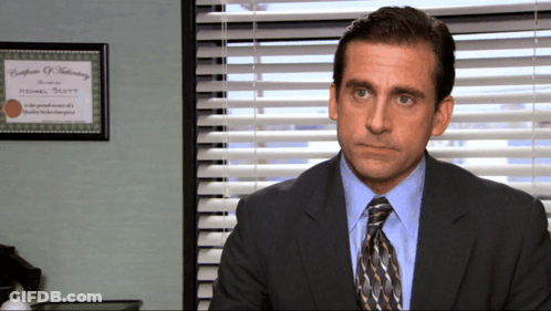 A Man In A Suit And Tie Sits In Front Of A Window With Blinds And A Certificate Of Training GIF