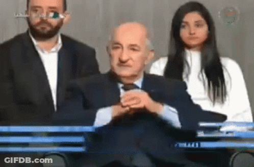 A Man In A Suit And Tie Sits In Front Of A Woman And A Man In A Suit GIF