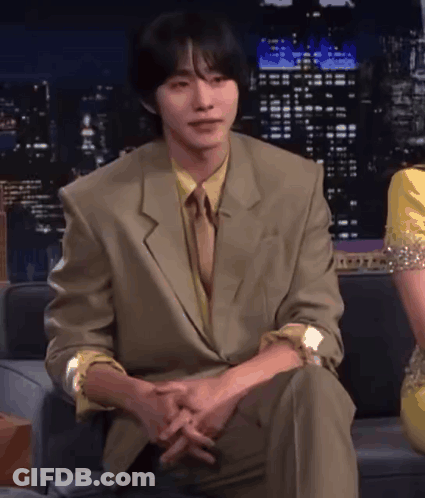 A Man In A Suit And Tie Sits On A Couch With His Hands Folded GIF