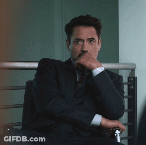 A Man In A Suit And Tie Sits With His Hand On His Chin GIF