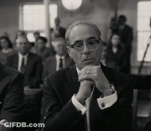 A Man In A Suit And Tie Sits With His Hands Folded In Front Of Him GIF