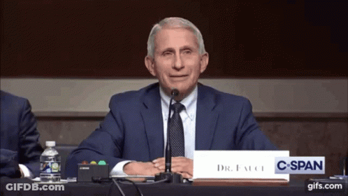 A Man In A Suit And Tie Sitting At A Table With A Sign That Says Dr. Fauci GIF