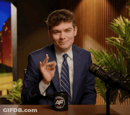 A Man In A Suit And Tie Sitting In Front Of A Microphone With A Mug That Says Avenue Af On It GIF