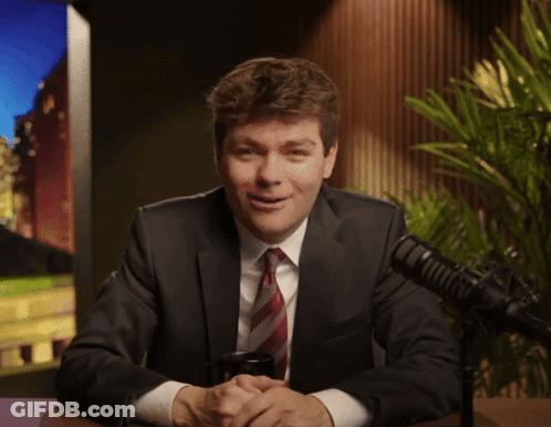 A Man In A Suit And Tie Smiles In Front Of A Microphone GIF