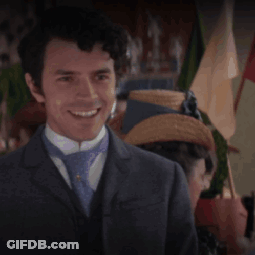 A Man In A Suit And Tie Smiles In Front Of A Woman In A Straw Hat GIF