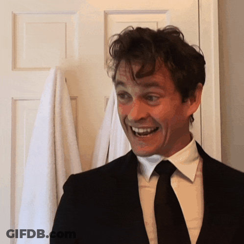 A Man In A Suit And Tie Smiles While Standing In Front Of A Door GIF