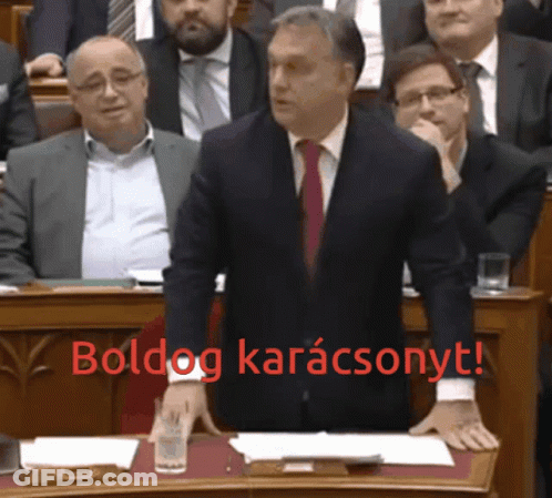 A Man In A Suit And Tie Stands At A Podium With The Words Boldog Karacsonyt In Red Behind Him GIF