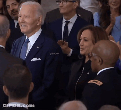 A Man In A Suit And Tie Stands In A Crowd With The Word Warbler On The Bottom Left GIF