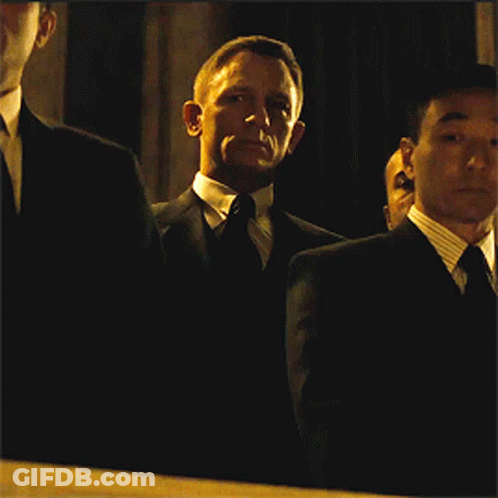 A Man In A Suit And Tie Stands In A Dark Room With Other Men GIF
