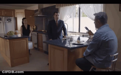 A Man In A Suit And Tie Stands In A Kitchen Talking To A Woman GIF