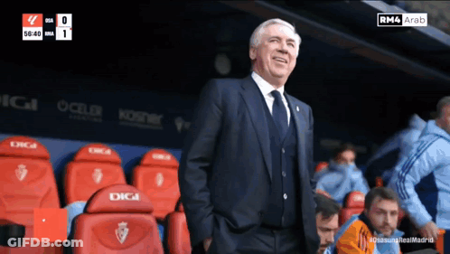 A Man In A Suit And Tie Stands In A Stadium With Red Seats That Say Digi GIF