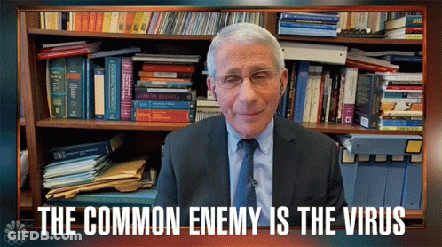 A Man In A Suit And Tie Stands In Front Of A Bookshelf With The Words " The Common Enemy Is The Virus " GIF
