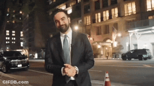 A Man In A Suit And Tie Stands In Front Of A Building At Night GIF