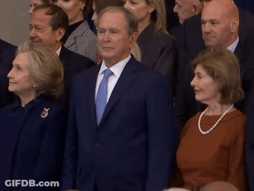 A Man In A Suit And Tie Stands In Front Of A Crowd Of People GIF