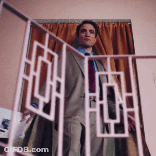 A Man In A Suit And Tie Stands In Front Of A Curtain GIF