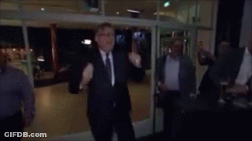 A Man In A Suit And Tie Stands In Front Of A Group Of People And Says " Voetjes Van De Vloer " In Yellow Letters GIF