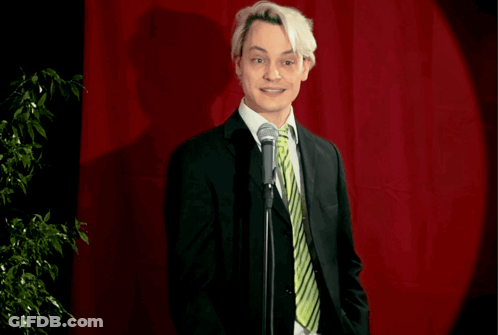 A Man In A Suit And Tie Stands In Front Of A Microphone With The Words " A Bit Desperate " Below Him GIF
