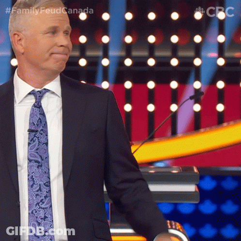 A Man In A Suit And Tie Stands In Front Of A Podium With The Words Family Feud Canada Written Above Him GIF