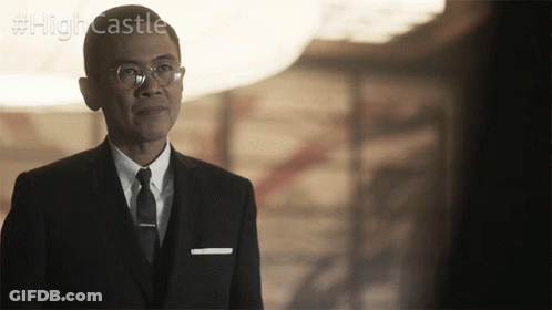 A Man In A Suit And Tie Stands In Front Of A Sign That Says High Castle GIF