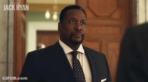 A Man In A Suit And Tie Stands In Front Of A Sign That Says " Jack Ryan " GIF