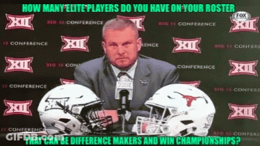 A Man In A Suit And Tie Stands In Front Of Two Football Helmets With Xii On Them GIF