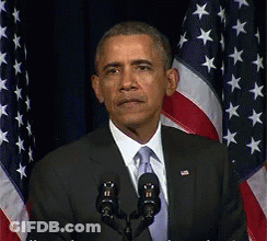 A Man In A Suit And Tie Stands In Front Of Two Microphones In Front Of An American Flag GIF