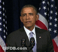 A Man In A Suit And Tie Stands In Front Of Two Microphones In Front Of An American Flag GIF