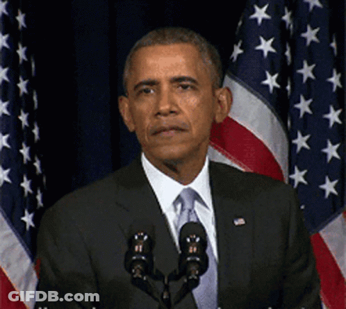 A Man In A Suit And Tie Stands In Front Of Two Microphones In Front Of An American Flag GIF