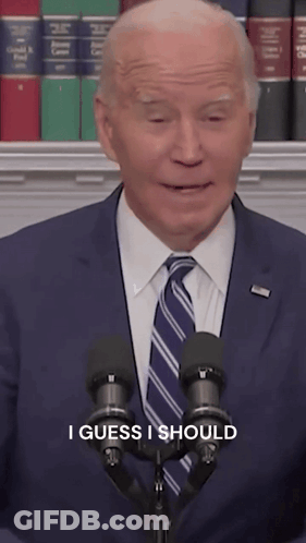 A Man In A Suit And Tie Stands In Front Of Two Microphones With The Words I Guess I Should Written Below Him GIF