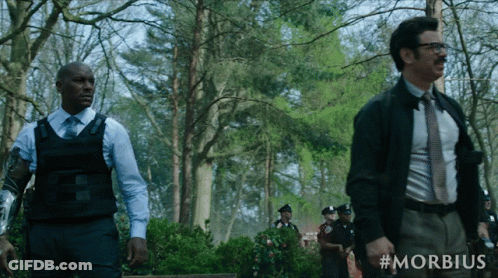 A Man In A Suit And Tie Stands Next To A Man In A Bullet Proof Vest In A Forest GIF