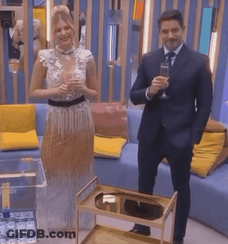 A Man In A Suit And Tie Stands Next To A Woman In A Dress Holding A Glass Of Champagne GIF