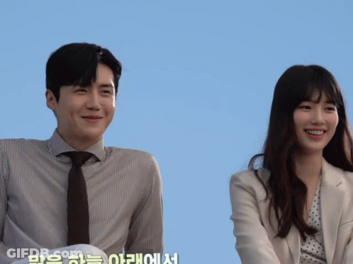 A Man In A Suit And Tie Stands Next To A Woman In A Polka Dot Jacket GIF
