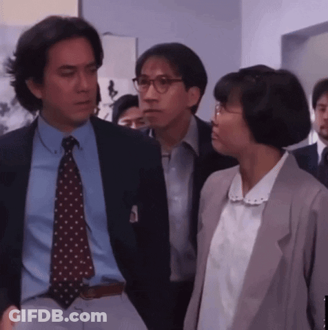 A Man In A Suit And Tie Stands Next To A Woman With Glasses GIF
