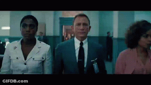 A Man In A Suit And Tie Stands Next To Two Women In A Hallway GIF