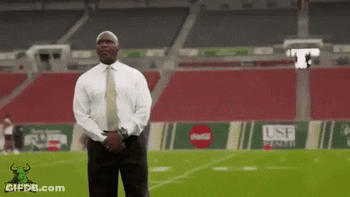 A Man In A Suit And Tie Stands On A Football Field With A Bullpen Logo On The Bottom GIF