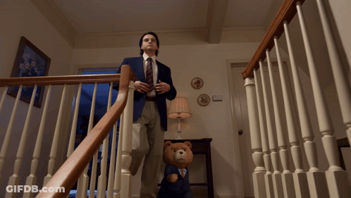 A Man In A Suit And Tie Stands On The Stairs Next To A Teddy Bear GIF