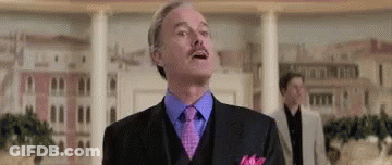 A Man In A Suit And Tie With A Mustache Is Standing In Front Of A Building . GIF
