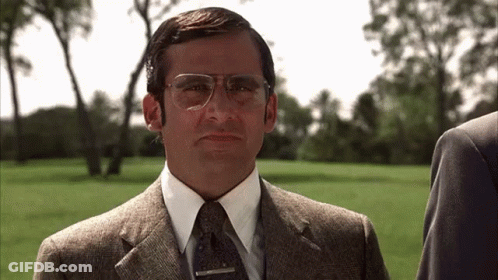 A Man In A Suit And Tie With Glasses Is Standing In A Grassy Field . GIF
