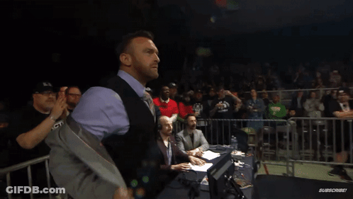 A Man In A Suit And Vest Stands In Front Of A Crowd With A Sign That Says ' Warriors ' On It GIF