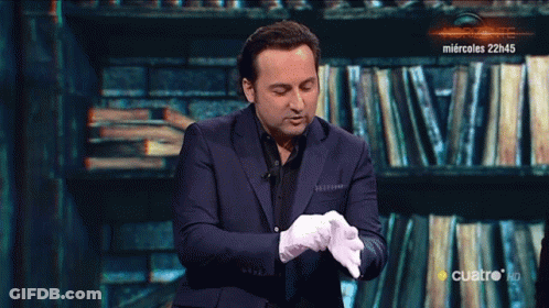 A Man In A Suit And White Gloves Stands In Front Of A Wall Of Books GIF