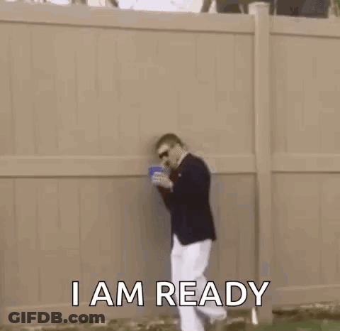 A Man In A Suit And White Pants Is Standing In The Grass With A Cup In His Hand . GIF