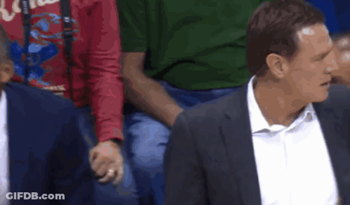 A Man In A Suit And White Shirt Is Sitting In A Stadium With Other Men . GIF