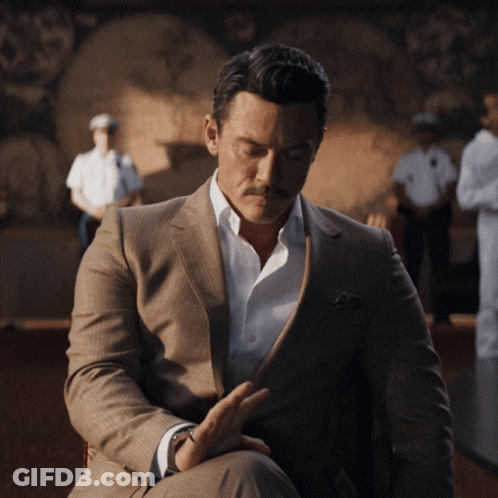 A Man In A Suit And White Shirt Is Sitting In Front Of A Painting GIF
