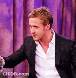 A Man In A Suit And White Shirt Is Sitting In Front Of A Purple Curtain And Holding A Glass Of Water . GIF