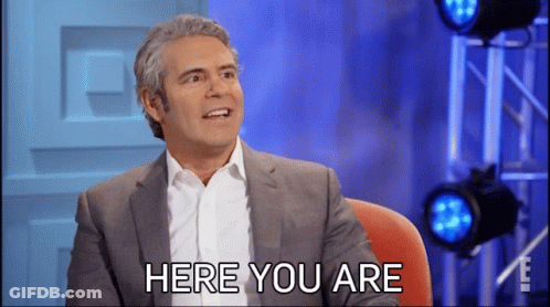 A Man In A Suit And White Shirt Is Sitting In Front Of A Sign That Says Here You Are GIF