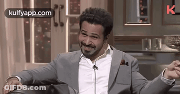 A Man In A Suit And White Shirt Is Sitting On A Couch With His Arms Outstretched And Smiling . GIF