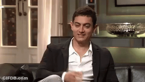 A Man In A Suit And White Shirt Is Sitting On A Couch With His Legs Crossed And Making A Funny Face . GIF